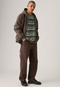Veste à capuche marron sur un t-shirt rayé vert et blanc, associée à un pantalon marron avec une poche latérale. Des chaussures noires complètent la tenue.
