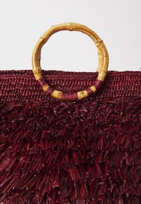 Burgundi szőtt táska, texturált bojt részletekkel, sötétebb rostba burkolt bambuszfogantyúval a kontraszt érdekében.