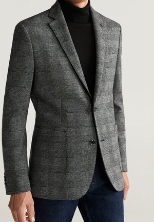 Mężczyzna w szarej, kratkowanej marynarce na czarnym golfie i ciemnoniebieskich dżinsach, stojący na tle jednolitego jasnego tła.