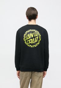 Černá mikina s velkým žlutým kruhovým motivem, na němž je nápis "SANTA CRUZ" a okolo okraje vzor šachovnice.