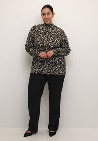 Blusa nera e beige a fantasia con collo alto, maniche lunghe e vestibilità ampia, abbinata a pantaloni neri a gamba dritta e scarpe con punta.