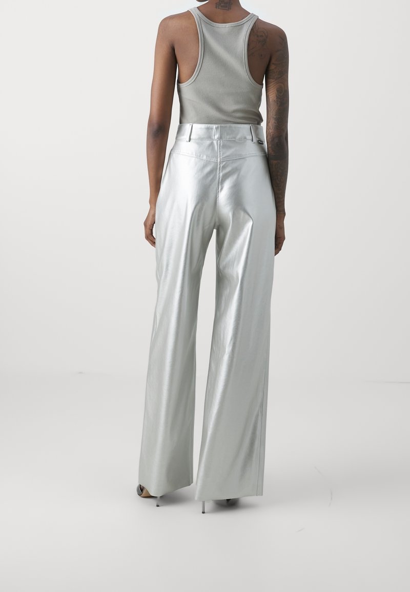 Personne portant un pantalon argenté à jambes larges et un haut gris sans manches, debout dos à nous sur fond blanc uni.