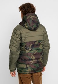 Pufferjakke med et grønt og brunt camouflage-mønster, elastiske ærmekanter og hætte. Glat, polstret stof med vandrette syninger.