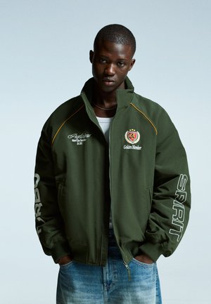 Green bomber jacket with a high collar, featuring embroidered graphics and a white "SPIRIT" print on the sleeve, paired with light blue jeans.