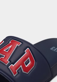 Sandale à semelle plate bleu marine avec logo "GAP" en relief rouge et blanc sur la bride, montrant une partie de la semelle intérieure avec le texte "GAP".