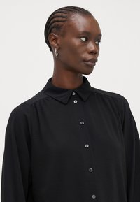 Blusa preta com colarinho e fecho à frente, feita de um tecido leve que apresenta uma superfície ligeiramente texturada e um detalhe subtil de pregas.