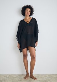 Čierny priesvitný kaftan s výstrihom do V, vo voľnom strihu a bočnými rozparkami. Doplnkovaný tenkými popruhmi. Nosený cez čierne plavky, naboso na textúrovanom podlahe.
