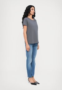 Grijs t-shirt met korte mouwen, ronde hals, gecombineerd met lichtblauwe jeans en zwarte spitse hakken. Glad materiaal, relaxte fit, zijaanzicht.