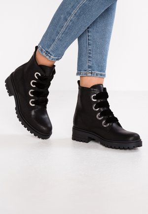 Bottines en cuir noir avec des lacets épais et des œillets argentés portées avec un jean skinny bleu court sur un sol blanc.