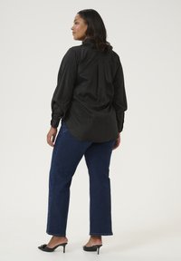 Blusa de satén negra con mangas largas, puño abotonado y pliegue en la parte trasera, combinada con jeans rectos de pierna oscura y tacones abiertos negros.