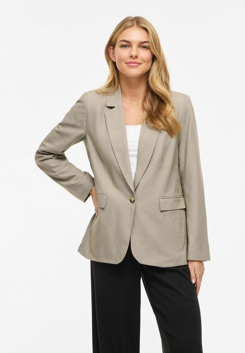 Femme aux longs cheveux blonds portant un blazer beige, un haut blanc et un pantalon noir, debout avec une main sur la hanche devant un fond blanc.