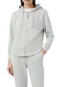 Jasnoszara bluza z kapturem na zamek w miękkim materiale, z kapturem ze sznurkiem, kieszenią na piersi oraz ściągaczami przy rękawach. Casualowy fason z zaokrąglonym dołem.