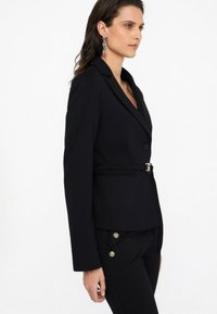 Blazer negro a medida con un diseño ajustado, cierre de un solo botón y un cinturón a juego. Tejido suave, solapas con muesca y mangas largas.