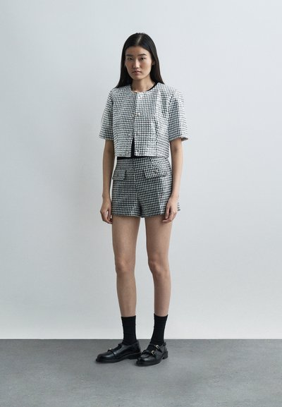 Jeune femme debout portant une veste à manches courtes en pied-de-poule noir et blanc et un short assorti, avec des chaussettes noires et des mocassins noirs sur un sol gris.