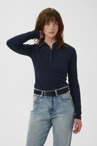 Polo de manga larga azul marino con cuello abotonado y textura acanalada, combinado con jeans de mezclilla de lavado claro y un cinturón negro con detalles plateados.