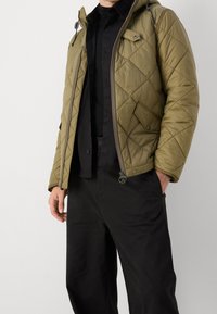 Olivgrüne gesteppte Jacke mit Kapuze, Reißverschluss und einer vorderen Tasche. Getragen über einem schwarzen Hemd und schwarzen Hosen, zeigt eine glatte Textur.