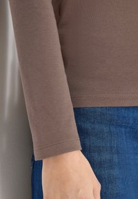 Hnědá košile s dlouhým rukávem, hladkou texturou, střihaným designem a prošitým lemem, spárovaná s modrými džínami z denimu ukazující hloubku látky.