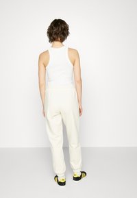 Top senza maniche bianco abbinato a pantaloni tuta crema. Sneakers nere e gialle presentano un logo e un design a strisce. Sfondo semplice.