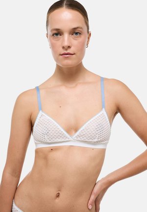Femme aux cheveux tirés en arrière portant un soutien-gorge en mesh blanc avec des bretelles bleu clair, posant avec une main sur la hanche devant un fond uni.