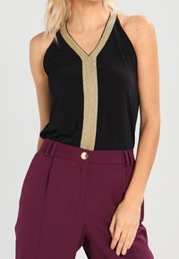 Svart ärmlös topp med V-ringning, prydd med guldglittrande kant längs mitten, tillsammans med höga burgundiska byxor.