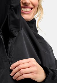 Schwarze wasserfeste Jacke mit glatter Textur, ausgestattet mit einem teilweisen Reißverschluss und verstellbaren Bündchen. Hände sind zu sehen, die den Reißverschluss anpassen.