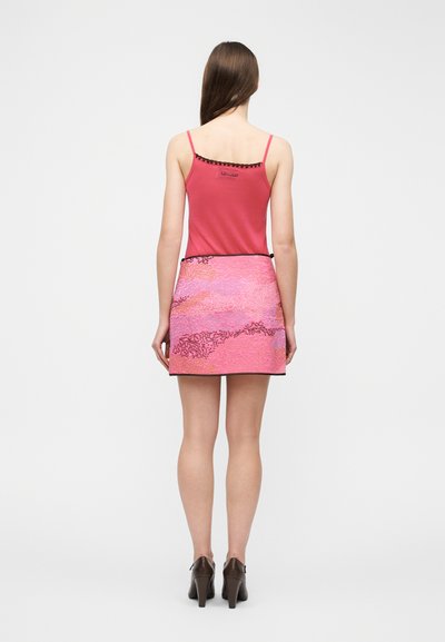 Γυναίκα που στέκεται με την πλάτη προς την κάμερα, φορώντας ένα ροζ αμάνικο τοπ, ροζ μίνι φούστα με σχέδια και καφέ ψηλοτάκουνα παπούτσια.