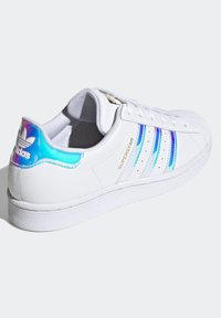 Vita sneakers med en slät syntetisk ovandel, tre iriserande ränder och en texturerad gummisula. Guldlogotyp på sidorna.