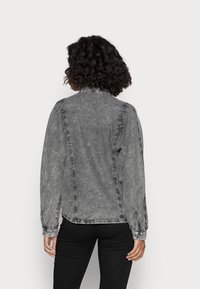 Kvinna med lockigt hår som bär en urblekt svart denimjacka och svarta byxor, stående vänd bortåt mot en neutral vit bakgrund.