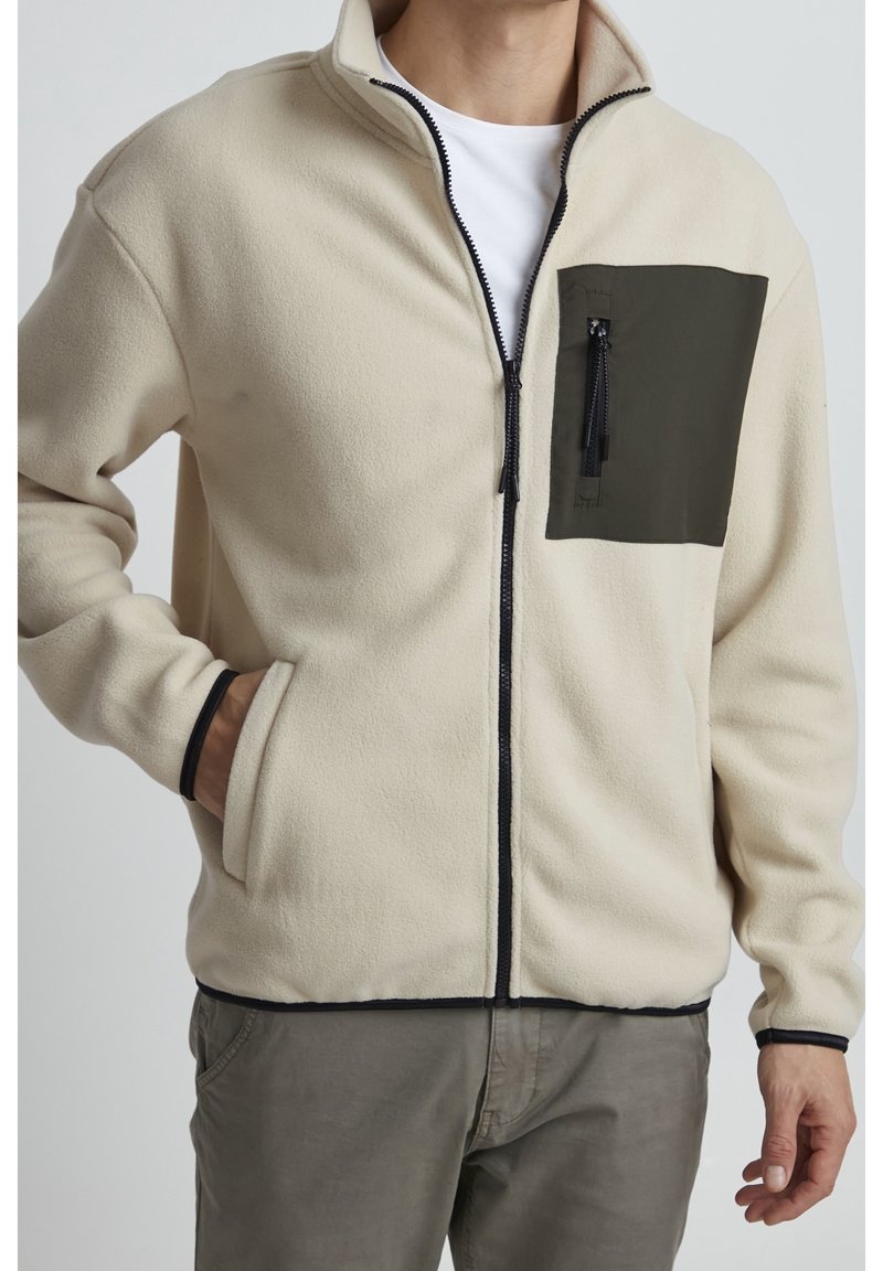 Veste zippée en polaire crème avec une poche poitrine verte. Elle présente un col haut, une fermeture éclair noire et un bord contrastant noir.