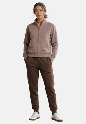 Femme debout les mains dans les poches, portant une veste zippée marron clair, un pantalon de jogging marron foncé et des baskets blanches sur fond blanc.