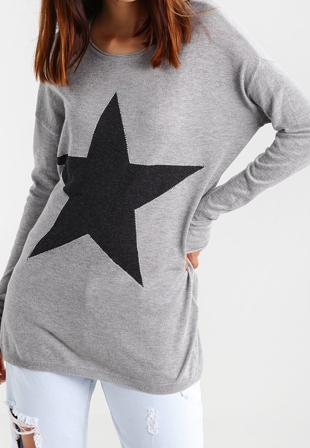 Szary sweter dzianinowy z dużym czarnym wzorem w gwiazdę z przodu. Posiada długie rękawy i luźny krój, zestawiony z jasnoniebieskimi, postarzanymi dżinsami.
