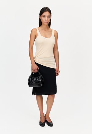 Femme portant un haut beige sans manches et une jupe noire, tenant un petit sac à main noir, debout sur un fond blanc uni.