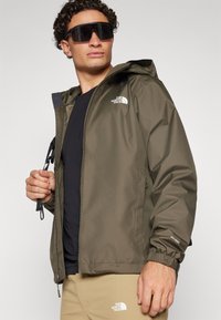 Jachetă impermeabilă de culoare verde măsliniu, cu glugă, manșete elastice și un logo pe piept. Purtați-o peste o bluză neagră și o pereche de pantaloni bej.