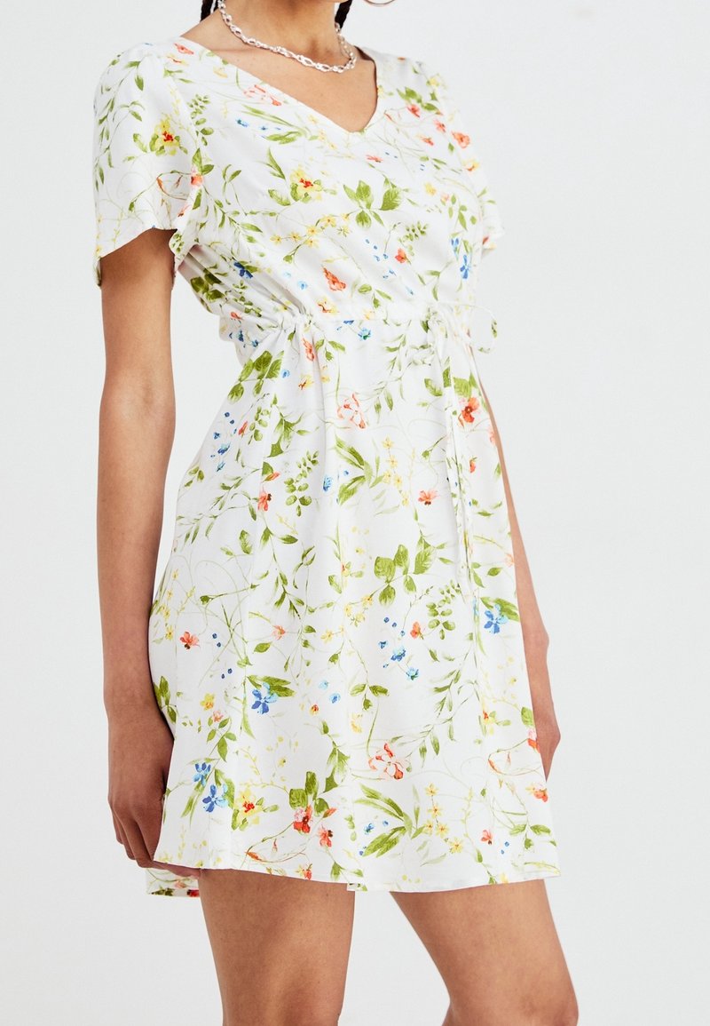 Robe fleurie en tissu léger, présentant un col en V, des manches courtes, une taille cintrée et des motifs floraux multicolores sur fond blanc.