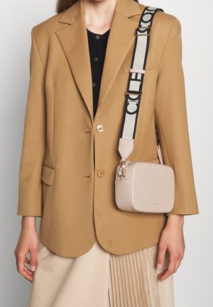 Persoon draagt een camelkleurig colbert over een zwart shirt en een beige geplooide rok, met een beige crossbodytas met een brede zwart-wit patroonriem.
