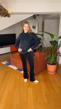 Pull noir en matière pelucheuse, pantalon évasé en velours noir et chaussures blanches. Décor intérieur avec un canapé brun texturé et une plante verte.