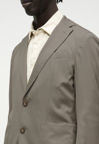 Άνδρας που φοράει ταμπά σακάκι με δύο κουμπιά πάνω από ένα εκρού πουκάμισο με γιακά, εμφανίζεται από τους ώμους ως το στήθος σε απλό φόντο.