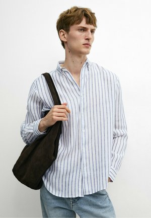 Jeune homme portant une chemise à rayures blanches et bleues avec boutons, jean bleu clair, portant un sac à épaule brun foncé, regardant sur le côté.