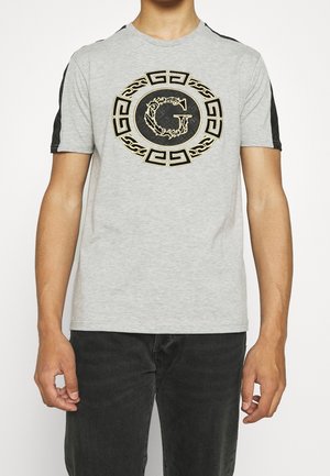 T-shirt en coton gris avec des accents noirs aux épaules, présentant un design graphique circulaire doré et noir avec la lettre "G" au centre.