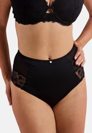 Femme portant un soutien-gorge en dentelle noire et une culotte taille haute noire avec des panneaux en résille florale, la main posée sur la hanche, sur un fond uni.