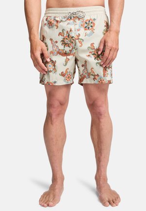 Uomo che indossa pantaloncini da bagno beige con motivi floreali arancioni e blu, in piedi a piedi nudi su uno sfondo bianco.