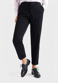 Pantalones negros ajustados con corte recto y dobladillo, combinados con zapatos de cuero verde oscuro con cordones y punta redonda.