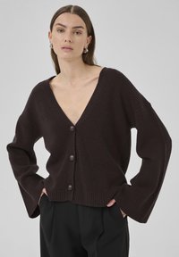 Cardigan en tricot marron avec un décolleté en V profond, des poignets côtelés proéminents et cinq grands boutons à l'avant. Texture douce, coupe décontractée.