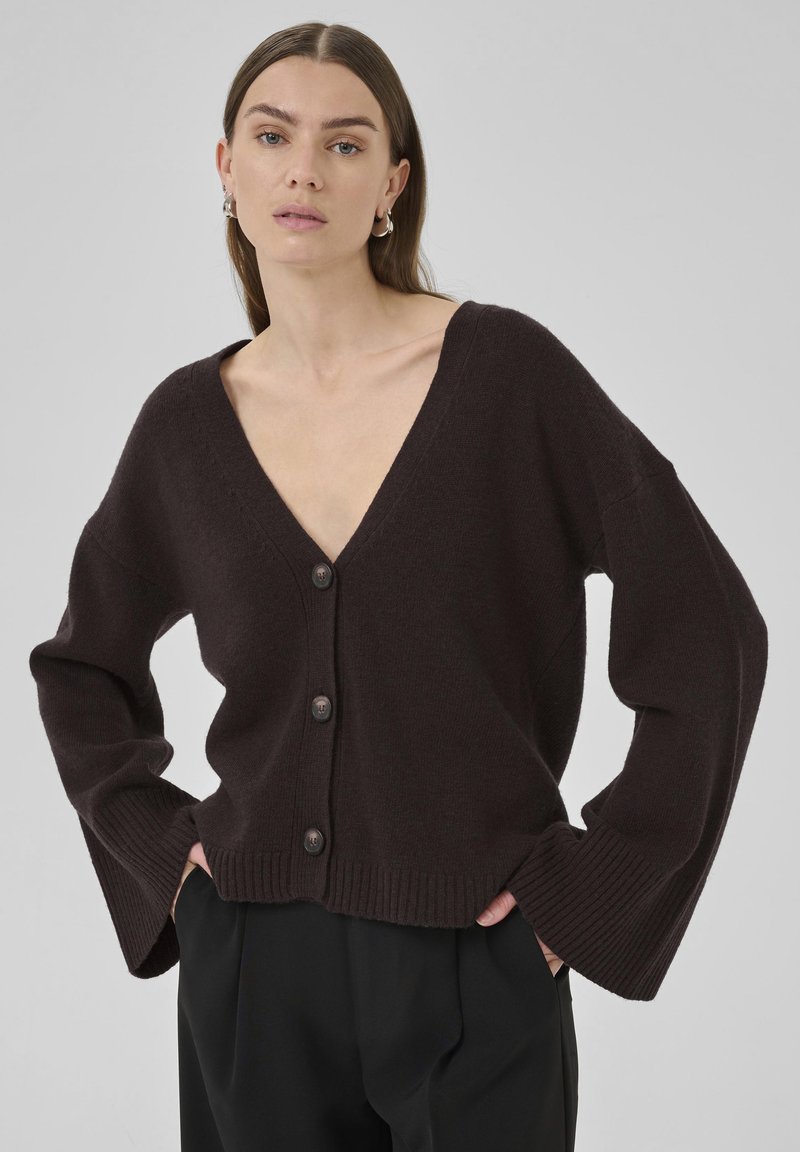 Cardigan en tricot marron avec un décolleté en V profond, des poignets côtelés proéminents et cinq grands boutons à l'avant. Texture douce, coupe décontractée.