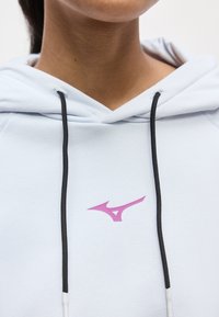 Κοντινή λήψη ενός ατόμου που φοράει λευκό φούτερ με μαύρα κορδόνια και ένα μικρό ροζ λογότυπο Mizuno στο στήθος.