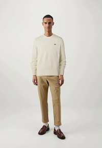 Kremfarget strikket genser med ribbestrikkede mansjetter, kombinert med beige chinos og mørkebrune loafers. Viser en diskret logo på brystet.