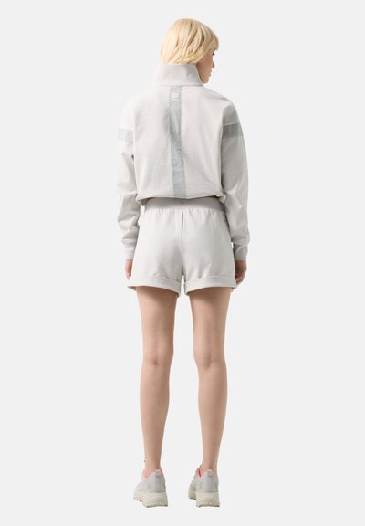 Personne blonde portant une veste blanche à col haut avec des bandes réfléchissantes et un short assorti à ourlet roulé, debout devant un fond uni.