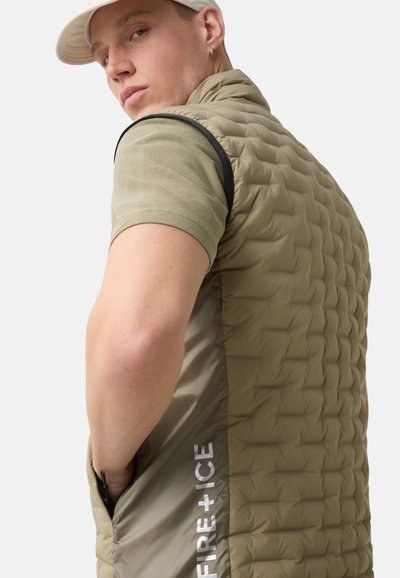 Padded olive green vest with a quilted texture, black accents, side pockets, and white logo detail on the lower edge. Designed for warmth and comfort.