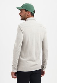 Hellgraues Langarmshirt mit Kragen, Schulternaht, schwarzen Knopfakzenten an den Ärmeln und einer glatten Textur. Getragen mit einer grünen Kappe.