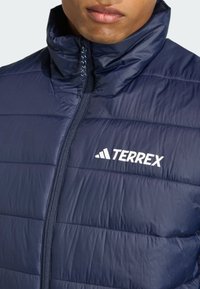 Marineblå pufferjakke med vertikal quiltet design, høj krave, frontlynlås og hvid "TERREX"-logo på brystet.
